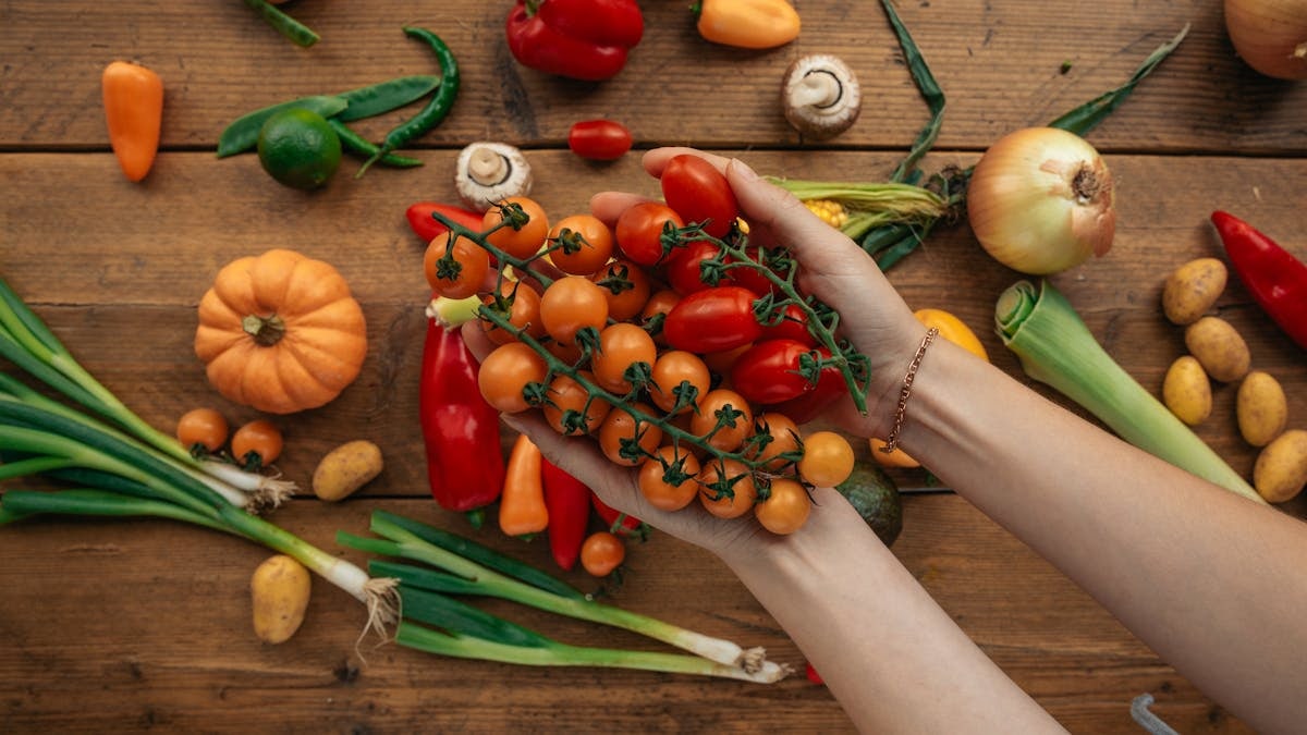 Hands holding cherry tomatoes surrounded by fresh vegetables on a rustic wooden surface.